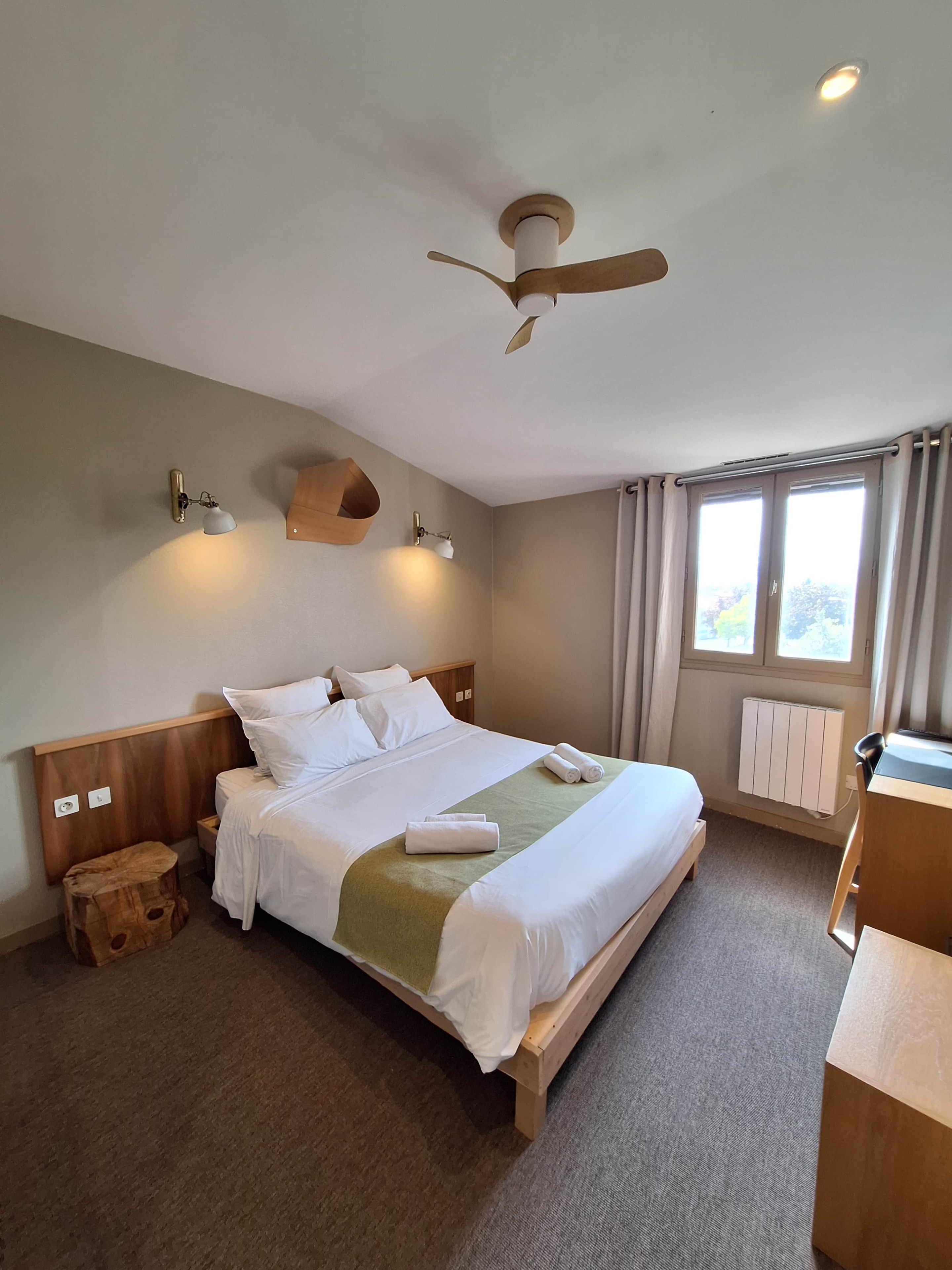 Une chambre d’hôtel avec un lit, un bureau, un ventilateur de plafond et une fenêtre avec des rideaux.