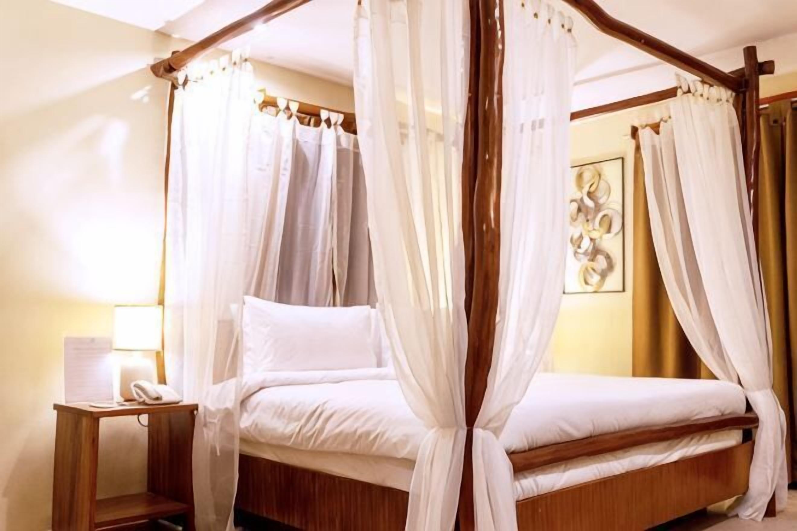 Una cama con dosel, cortinas translúcidas, una mesita de noche con lámpara y una obra de arte colgada en la pared.