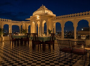 A checkered floor, a dining area with tables and chairs, and a view of the city at night. - Hotel Amar Kothi (Udaipur)