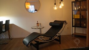 A modern lounge area with a reclining chair, a round mirror, a small table with a bottle, and a shelf with glassware. - Vacation apartment Dichter & Denker (Weimar)