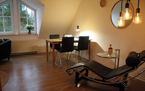 A modern loft bedroom with a sloped ceiling, a dining area with a table and chairs, a large mirror, and a lounge chair.