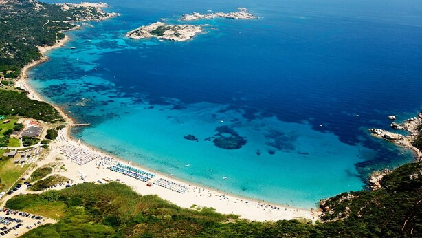 Vue aérienne d’une zone côtière avec une plage, des affleurements rocheux et des eaux d’un bleu limpide.