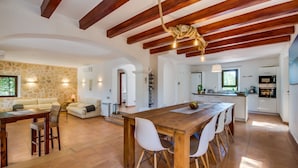 A spacious living area with a wooden dining table, white chairs, a stone wall, and a kitchen in the background. - Country House Finca Kiana with Pool, Garden & Wi-Fi (Sencelles)
