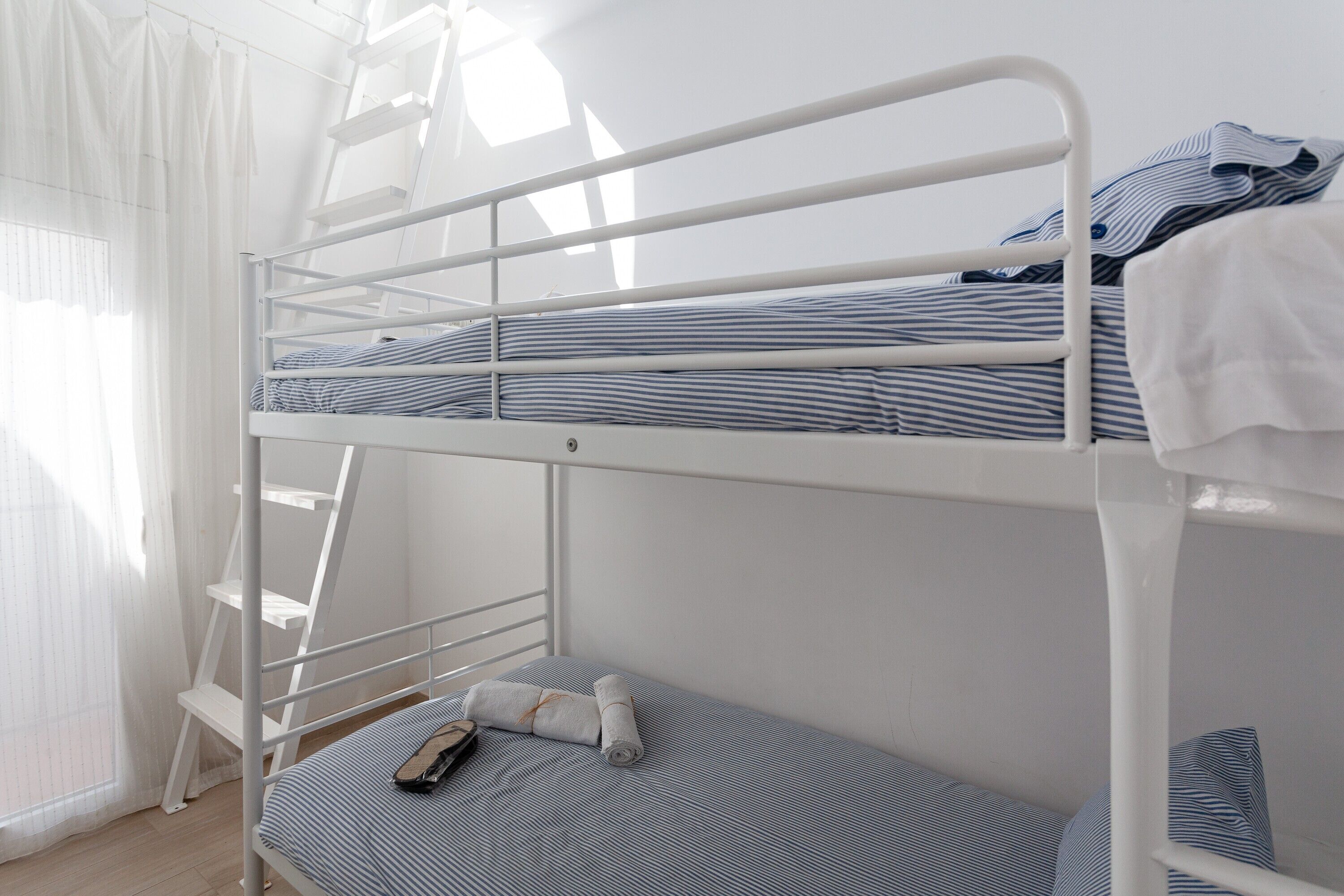 Un lit superposé avec une échelle, une petite table et une fenêtre ornée de rideaux légers.