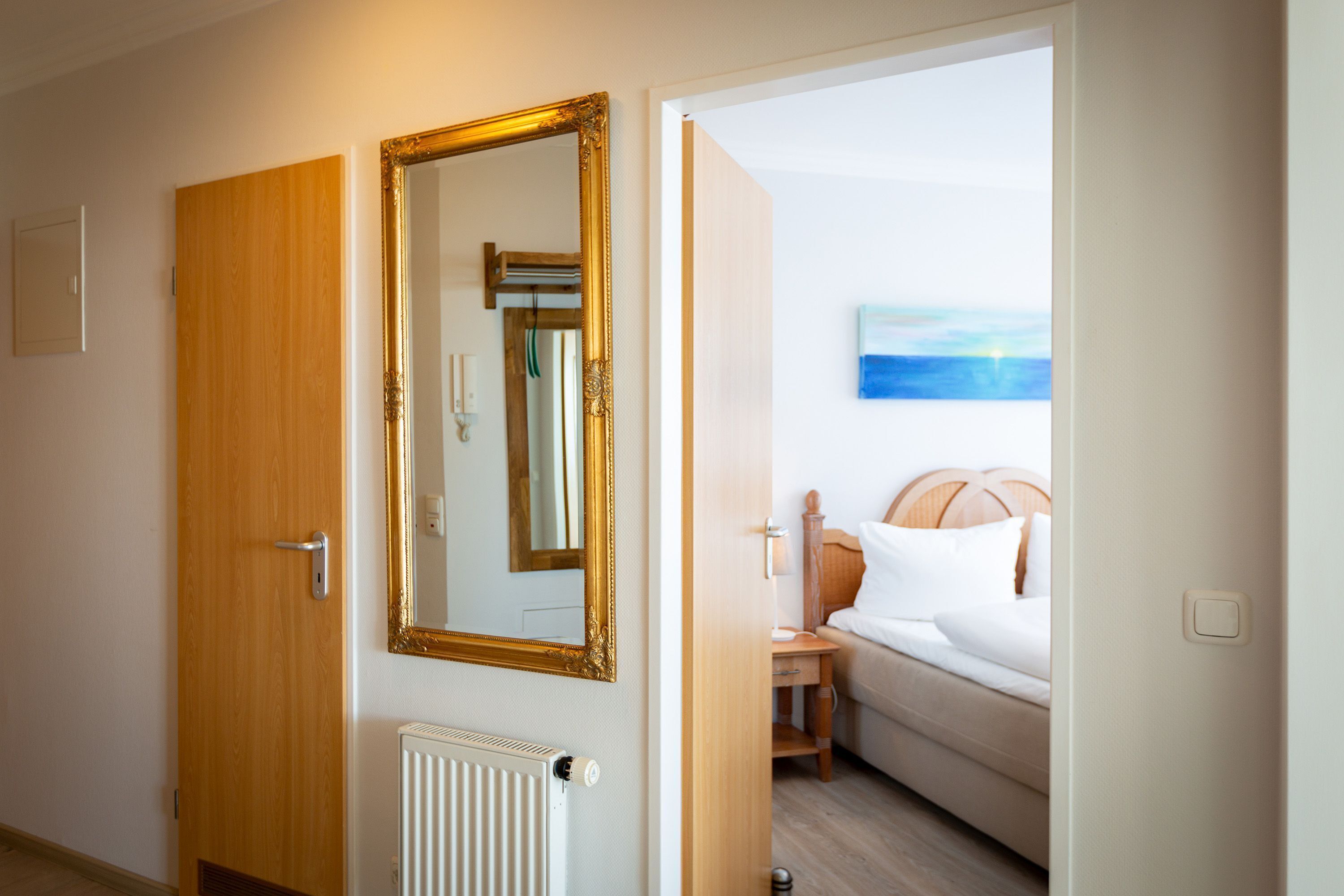 Quarto de hotel com porta de madeira, um espelho grande, radiador e cama com roupa de cama branca.