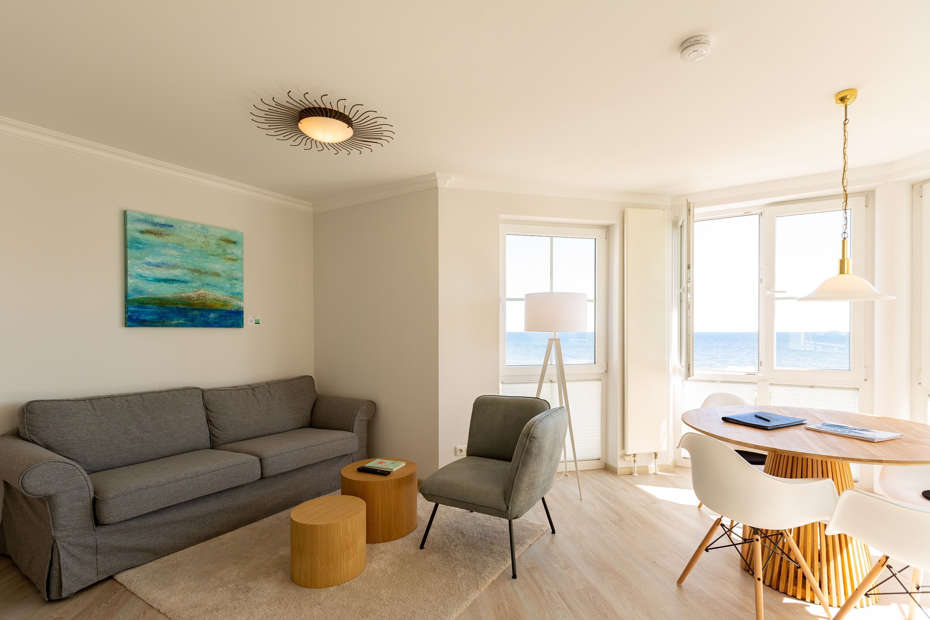 Una sala de estar moderna con un sofá gris, una mesita redonda, un sillón gris y una mesa de comedor de madera con sillas. Hay un cuadro en la pared y una ventana con vistas al mar.