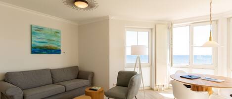 Una sala de estar moderna con un sofá gris, una mesita redonda, un sillón gris y una mesa de comedor de madera con sillas. Hay un cuadro en la pared y una ventana con vistas al mar.