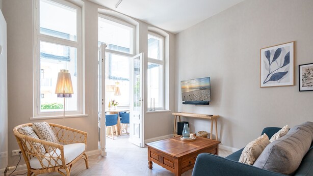 A spacious living room with a large window, a wooden coffee table, a blue sofa, and a wicker chair. - Villa Goodewind, App. 1.1 (Heringsdorf)