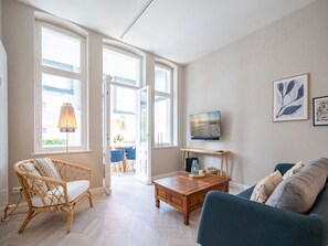 Uma sala de estar ampla com uma grande janela, uma mesa de centro de madeira, um sofá azul e uma poltrona de vime.