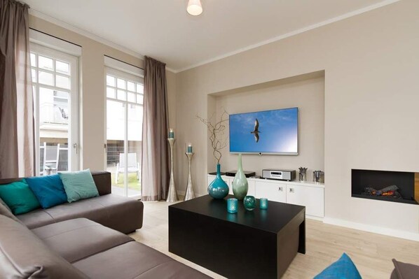 Ein modernes Wohnzimmer mit Sofa, Couchtisch, Fernseher, Kamin und großen Fenstern.