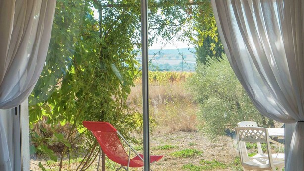 A room with a large window, a sofa with a patterned throw, a red chair, and a white chair outside. - Holiday Home in Murviel with French Garden (Murviel-Les-Beziers)
