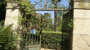 An ornate wrought iron gate with a decorative top, leading to a well-maintained garden area with trimmed hedges and palm trees. - Holiday Home in Murviel with French Garden (Murviel-Les-Beziers)