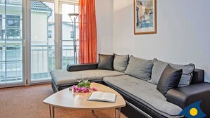 A modern living room with a sectional sofa, a small round table with a book and flowers, and a large window with blinds. - Villa Maria-Gabriele Whg. 09 //- - + Maria 09 (Loddin)