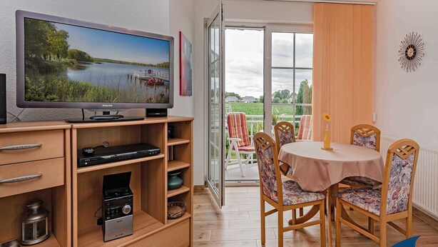 A living room with a television, wooden entertainment unit, and a dining area with a table and chairs. - Villa Maria-Gabriele Whg. 09 //- - + Maria 09 (Loddin)