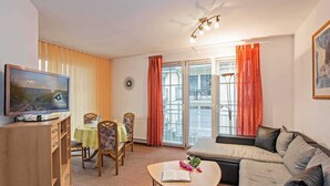 A living room with a television, a dining table with chairs, a sofa, and a coffee table with a book and flowers. - Villa Maria-Gabriele Whg. 09 //- - + Maria 09 (Loddin)