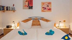 A hotel room with a wooden headboard, two white pillows, a bed with blue towels and white flowers, a bedside table with a lamp, a shelf with books, and two wall art pieces. - Villa Maria-Gabriele Whg. 09 //- - + Maria 09 (Loddin)
