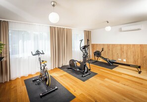 A home gym with exercise bikes and a rowing machine on black mats, wooden flooring, and large windows with curtains. - Citadines South Vienna (Vienna)