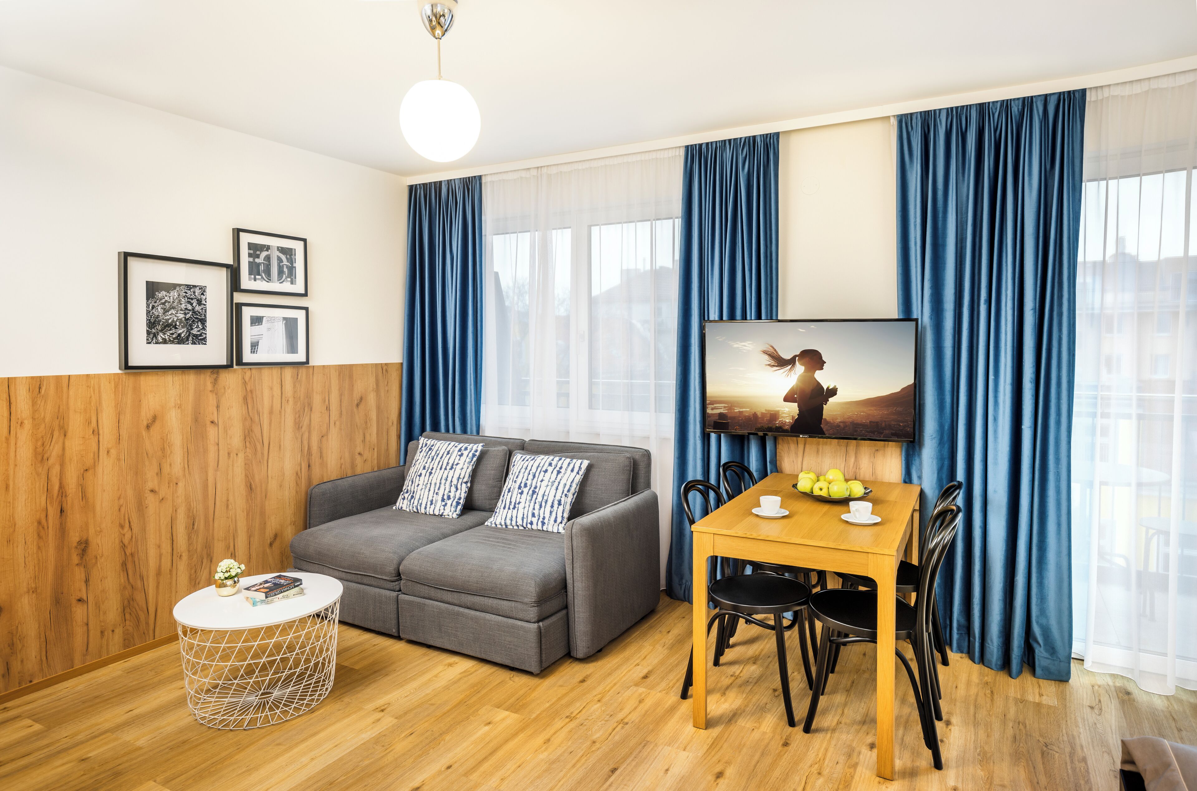 Una sala de estar moderna con un sofá gris, una mesa de comedor de madera con sillas, una mesa redonda de centro y un televisor montado en la pared.