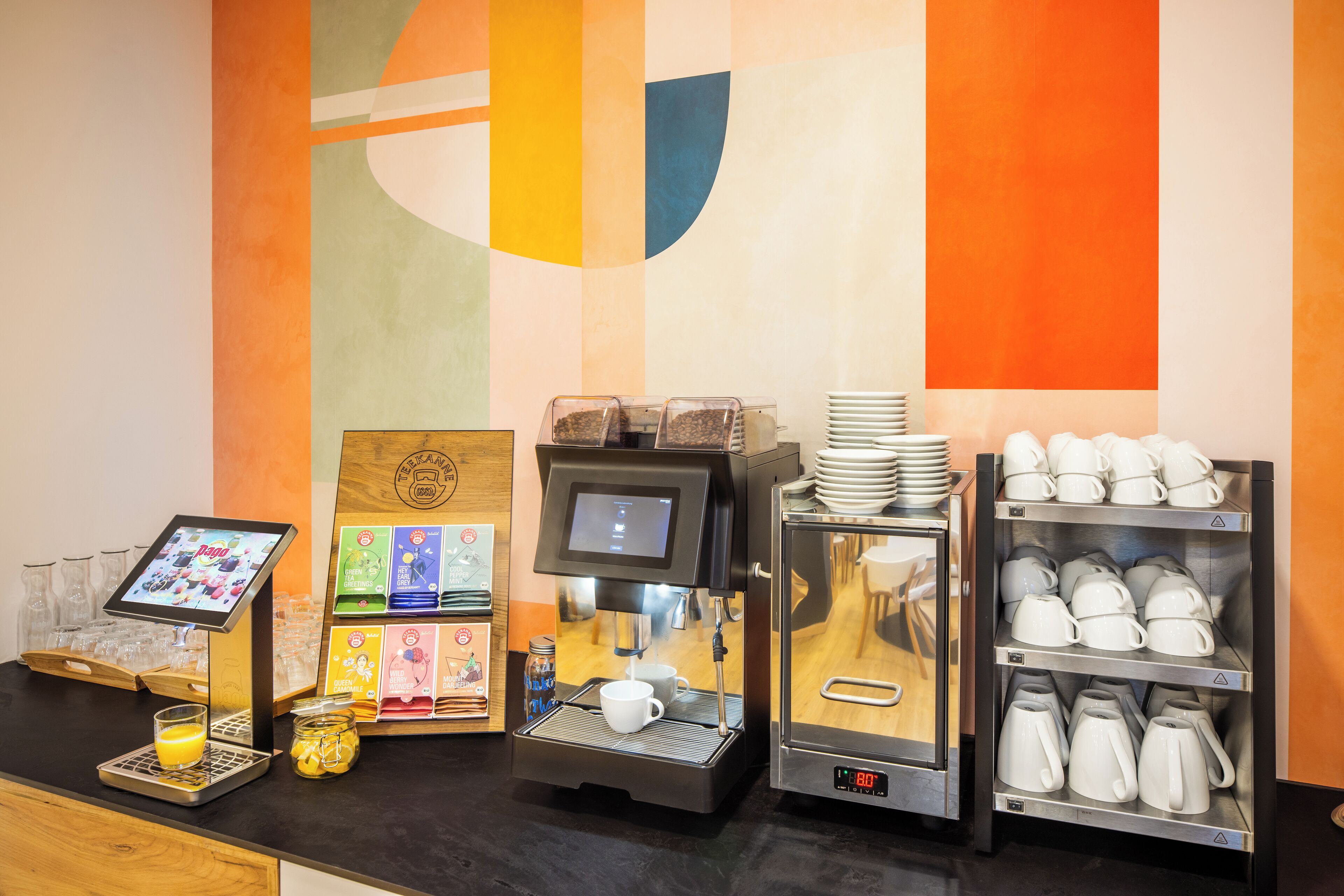 A coffee station with a digital ordering screen, a coffee machine, and a display of cups and mugs.