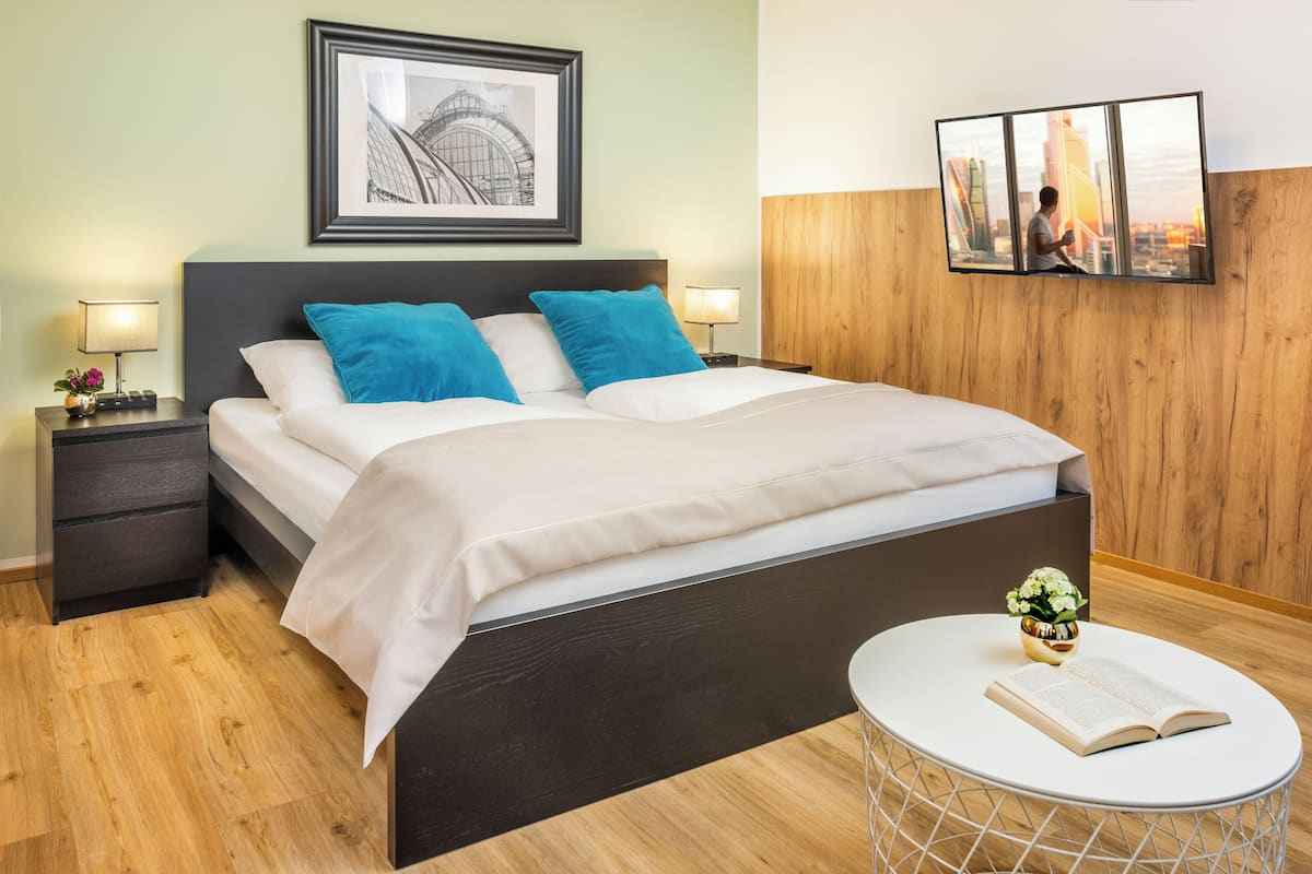 A modern hotel room with a large bed, two bedside tables with lamps, a round white table with a book and flowers, and a framed picture above the bed.