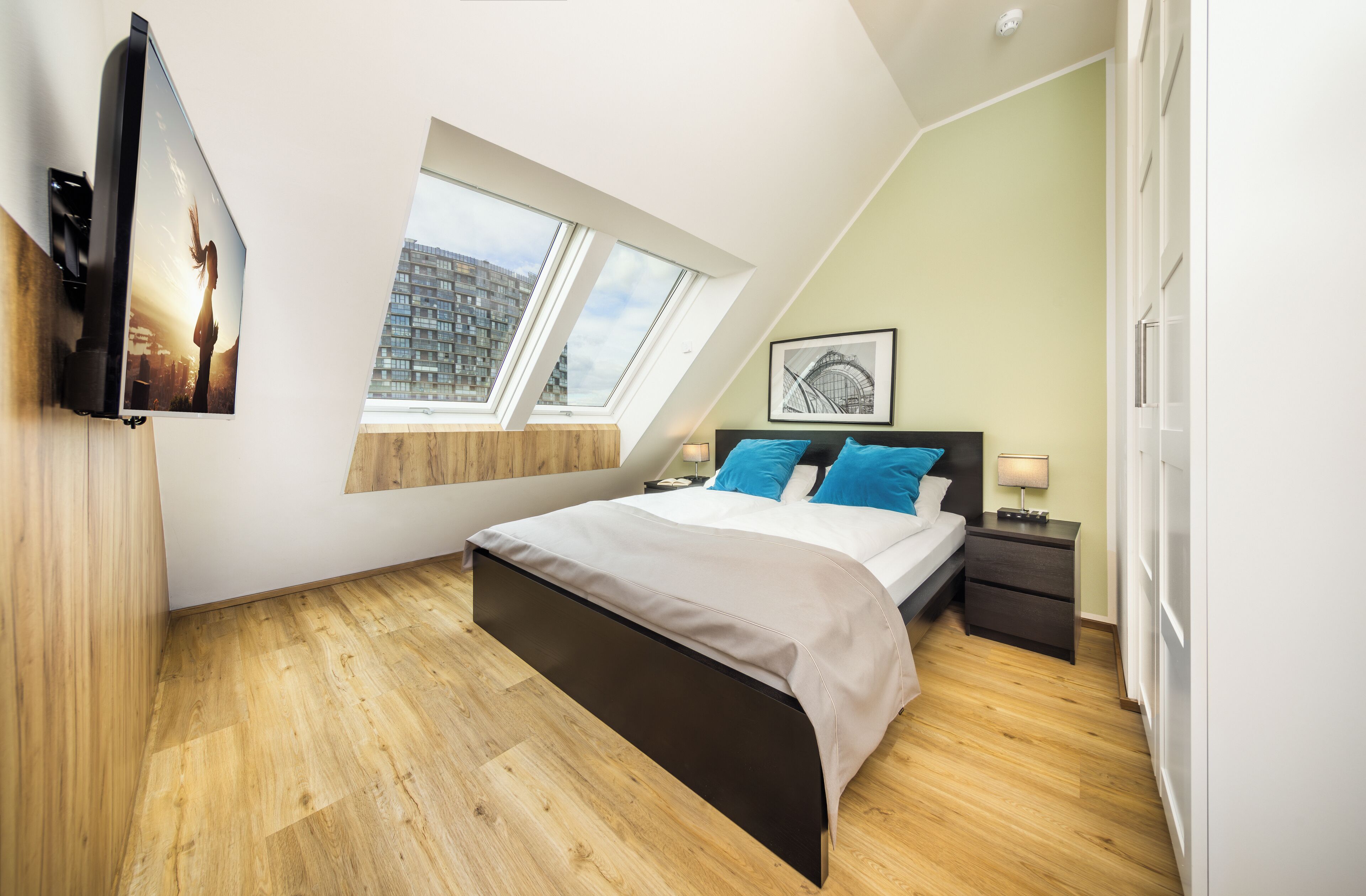 A bedroom with a bed, a nightstand, a TV mounted on the wall, a window, and a picture frame on the wall.