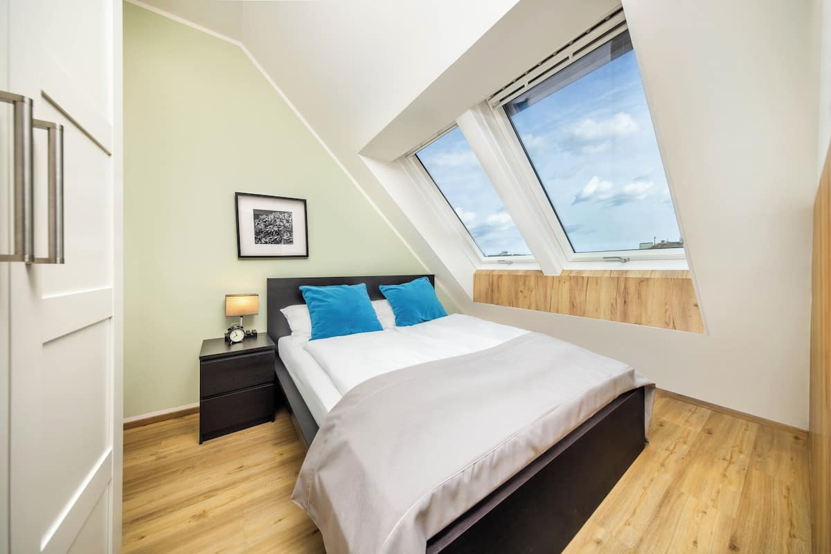 A bedroom with a skylight, a bed with blue pillows, a bedside table with a lamp, and a framed picture on the wall.