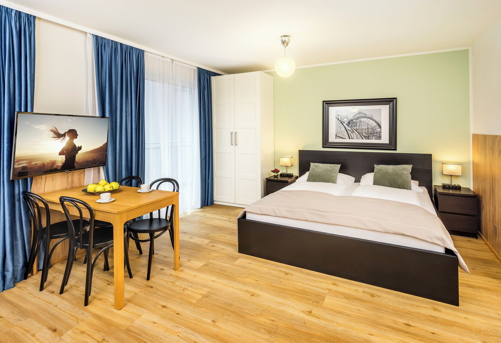 Hotellihuone, jossa on sänky, työpöytä tuoleineen, ruokapöytä hedelmillä ja seinällä taulu.