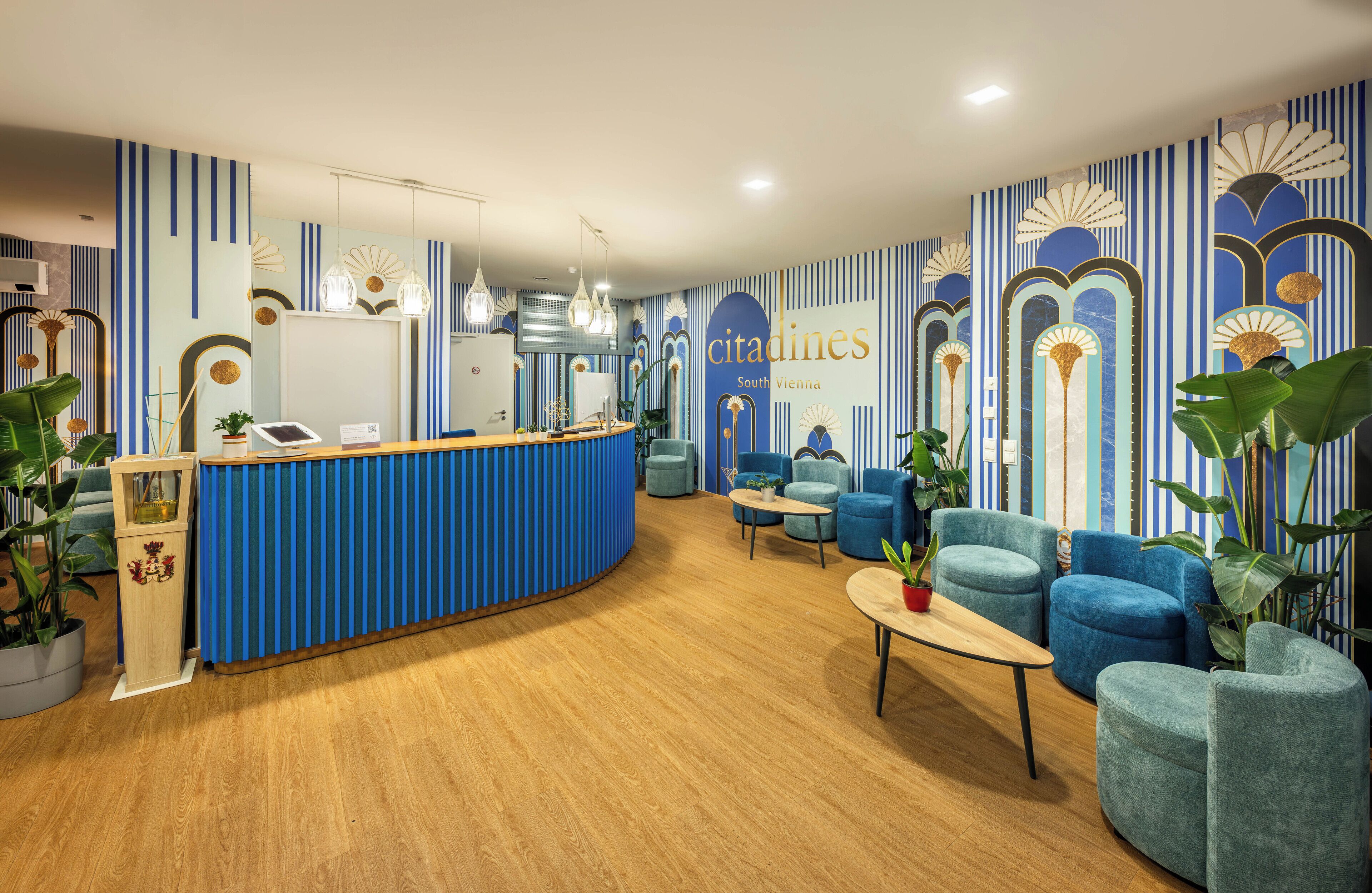 Un área de recepción con mostrador azul, asientos con plantas en macetas y decoración en la pared.