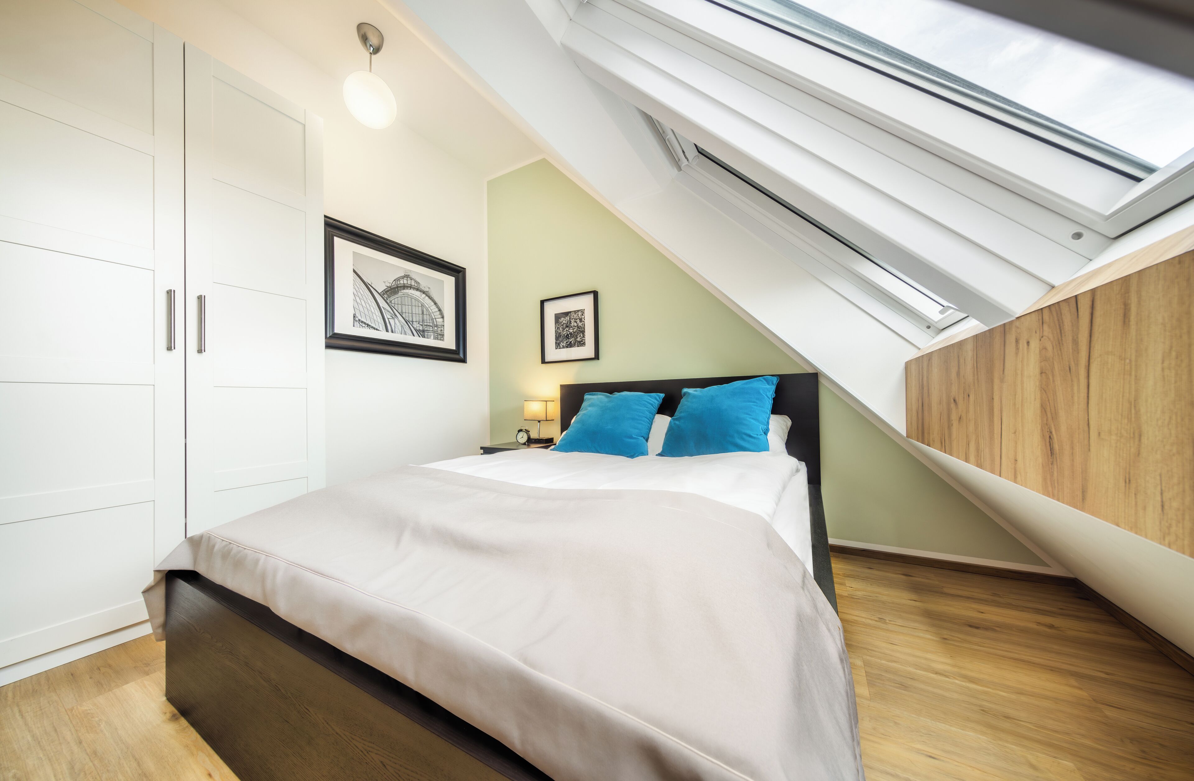 A bedroom with a skylight, a bed with blue pillows, a nightstand, and a framed picture on the wall.