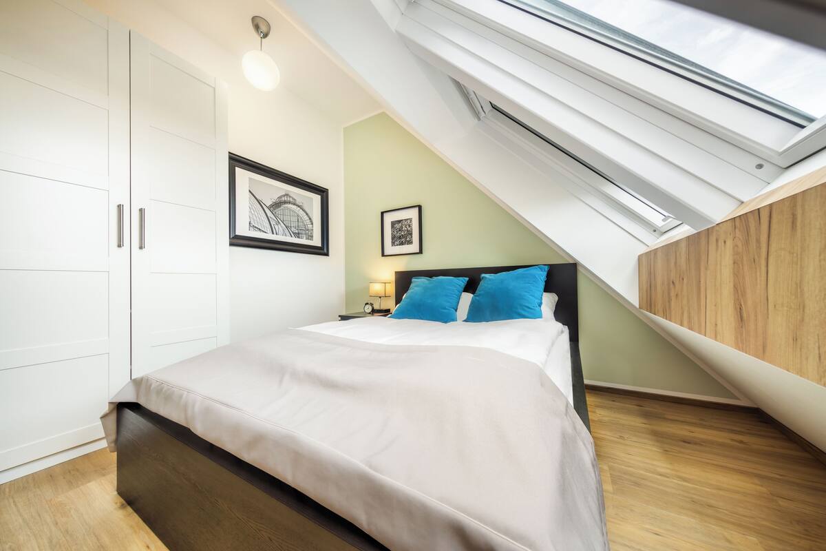 A bedroom with a skylight, a bed with blue pillows, a nightstand, and a framed picture on the wall.