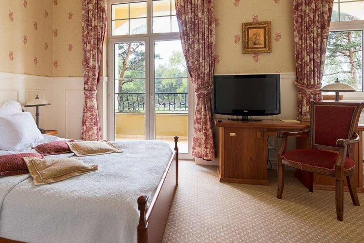 Ein Hotelzimmer mit einem großen Bett, einem Schreibtisch mit Fernseher, einem Sessel und einem Fenster mit Vorhängen.