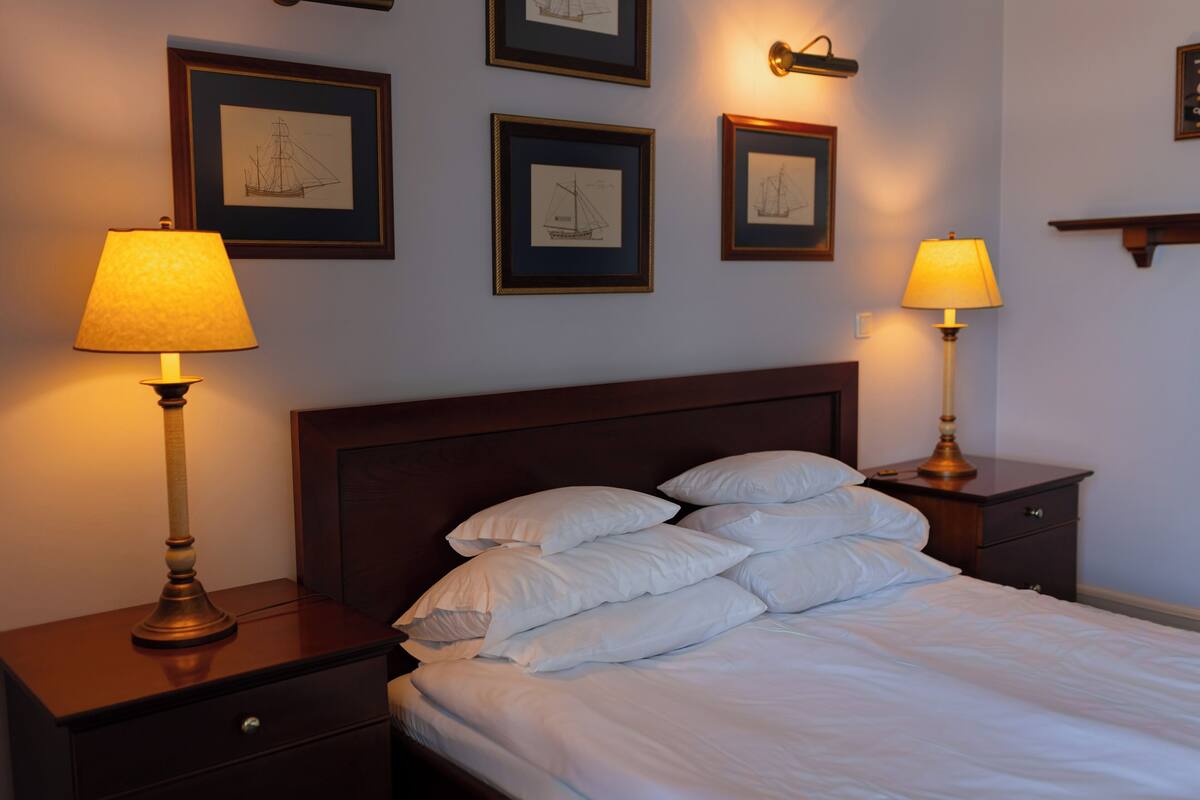 Ein ordentlich bezogenes Bett mit weißen Leinen, zwei Nachttische mit Lampen und gerahmte maritime Zeichnungen an der Wand.