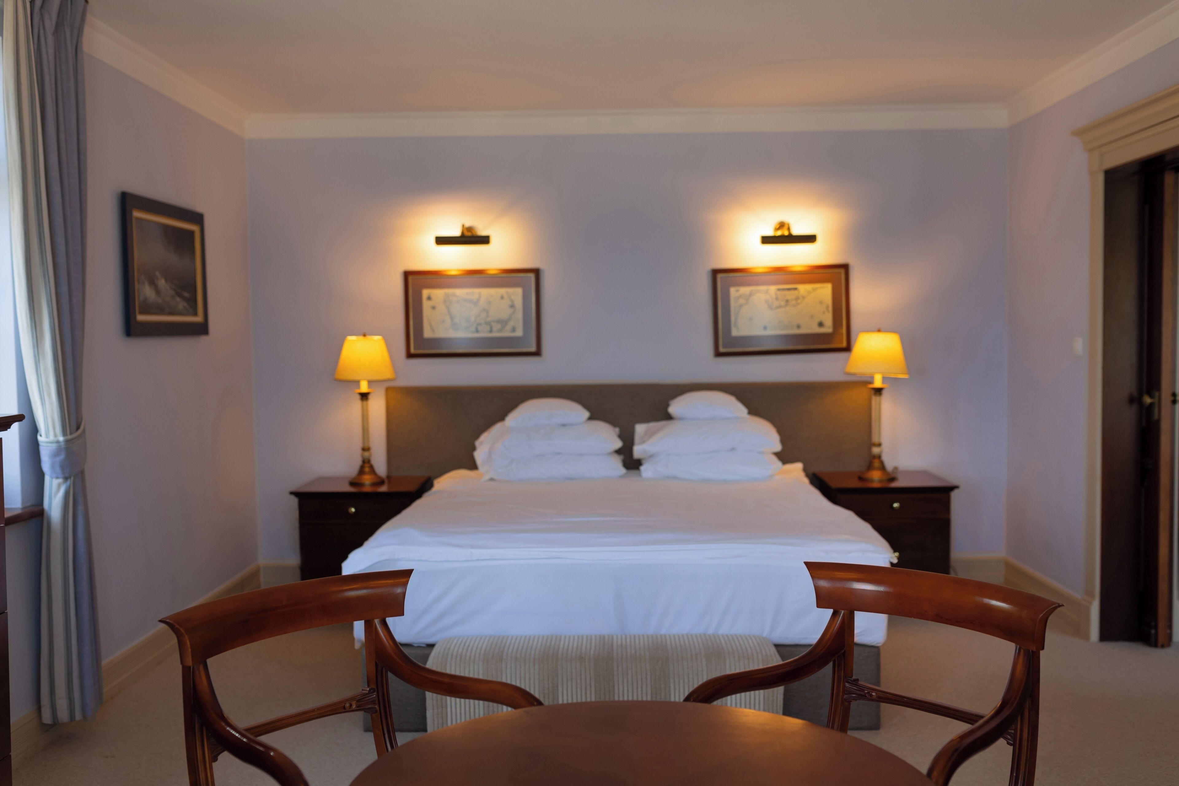 A hotel room with a bed, two bedside lamps, a framed picture on the wall, and a chair at the foot of the bed.