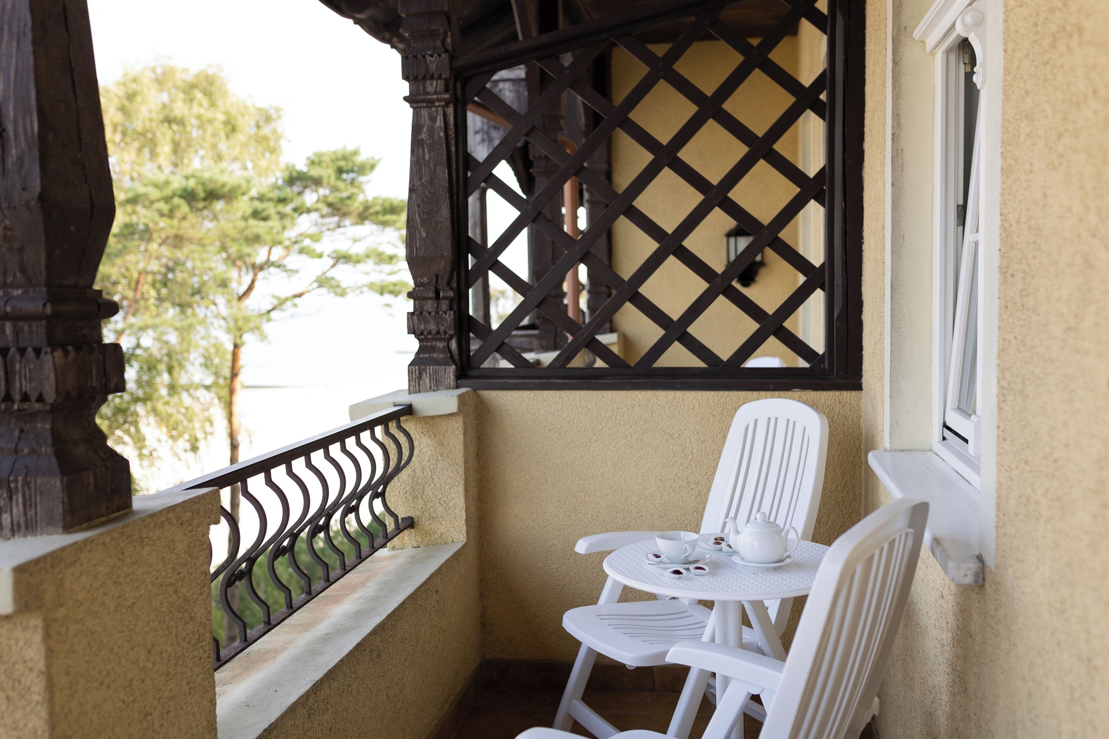 A small balcony with a white table and chairs, a decorative black lattice, and a view of trees.