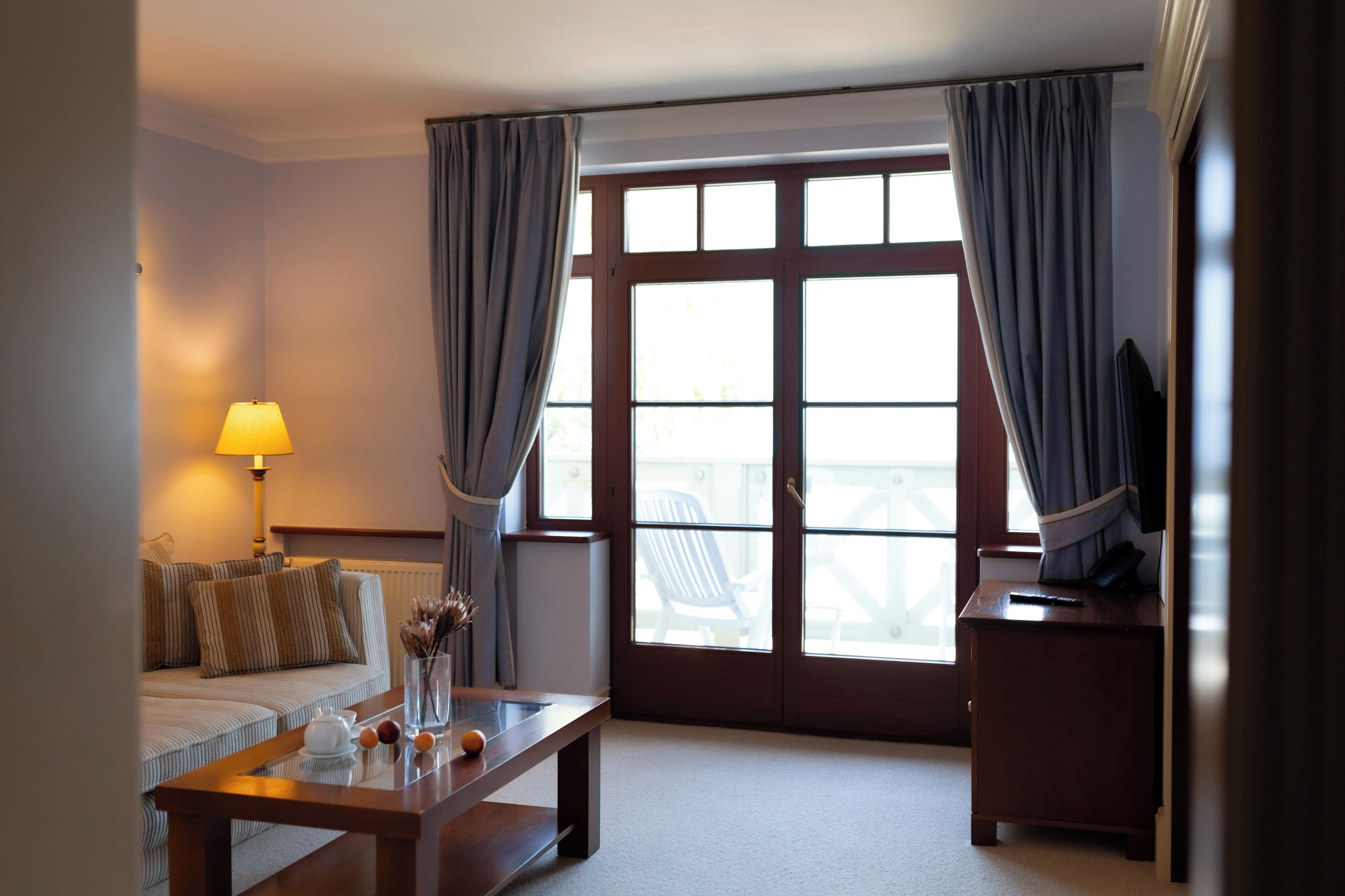 A hotel room with a sofa, a small table with a lamp, a TV on a stand, and a large window with curtains.