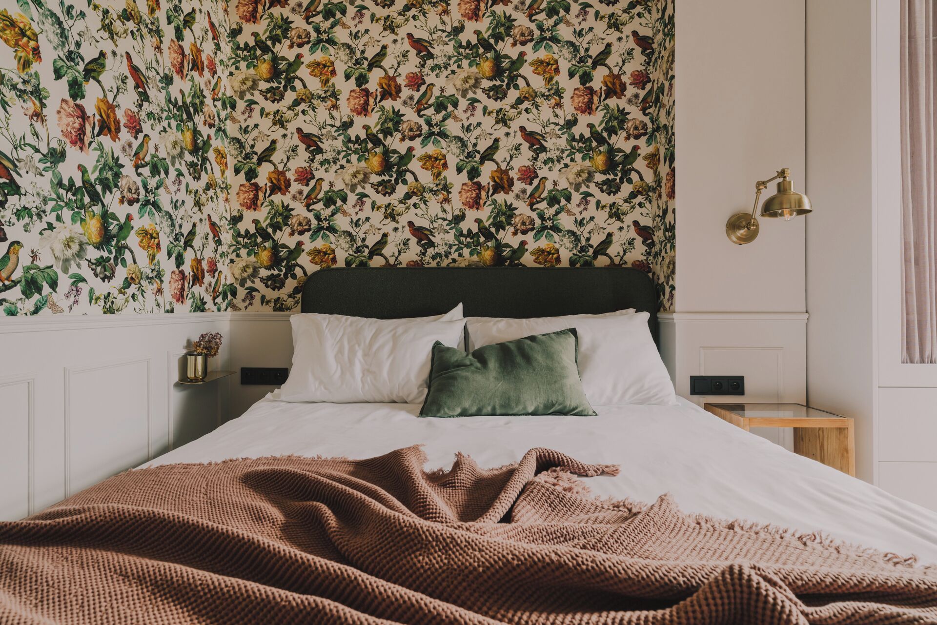 Une chambre avec un papier peint à motifs floraux, un lit recouvert de draps blancs et orné d’un coussin vert, une table de chevet en bois avec une lampe, et une fenêtre avec des rideaux.