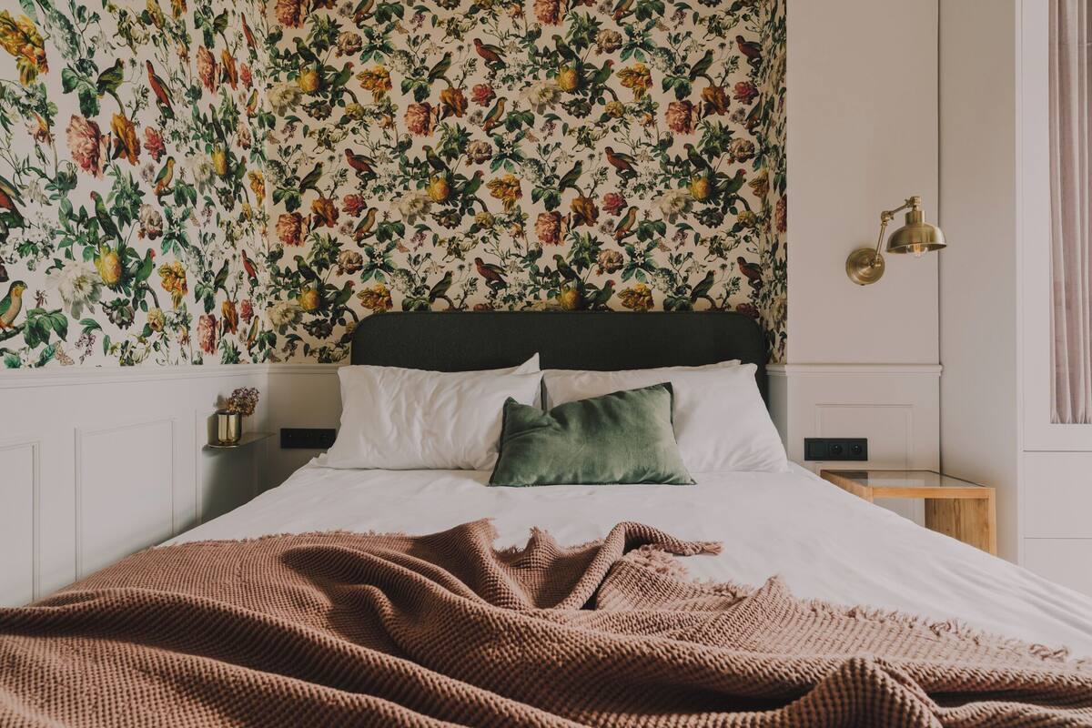 Ein Schlafzimmer mit floraler Tapete, einem Bett mit weißen Bettbezügen und einem grünen Kissen, einem hölzernen Nachttisch mit Lampe und einem Fenster mit Vorhängen.