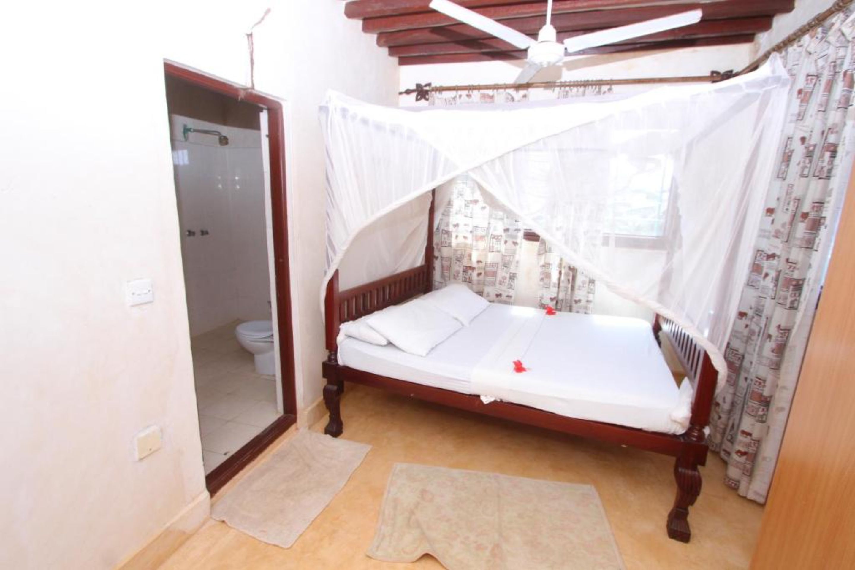 A bedroom with a wooden bed, a ceiling fan, a window with curtains, and a door leading to a bathroom.
