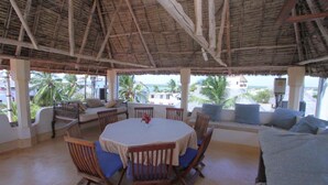 Un área de comedor al aire libre techada con una mesa redonda y sillas de madera, con vista a una playa con palmeras.