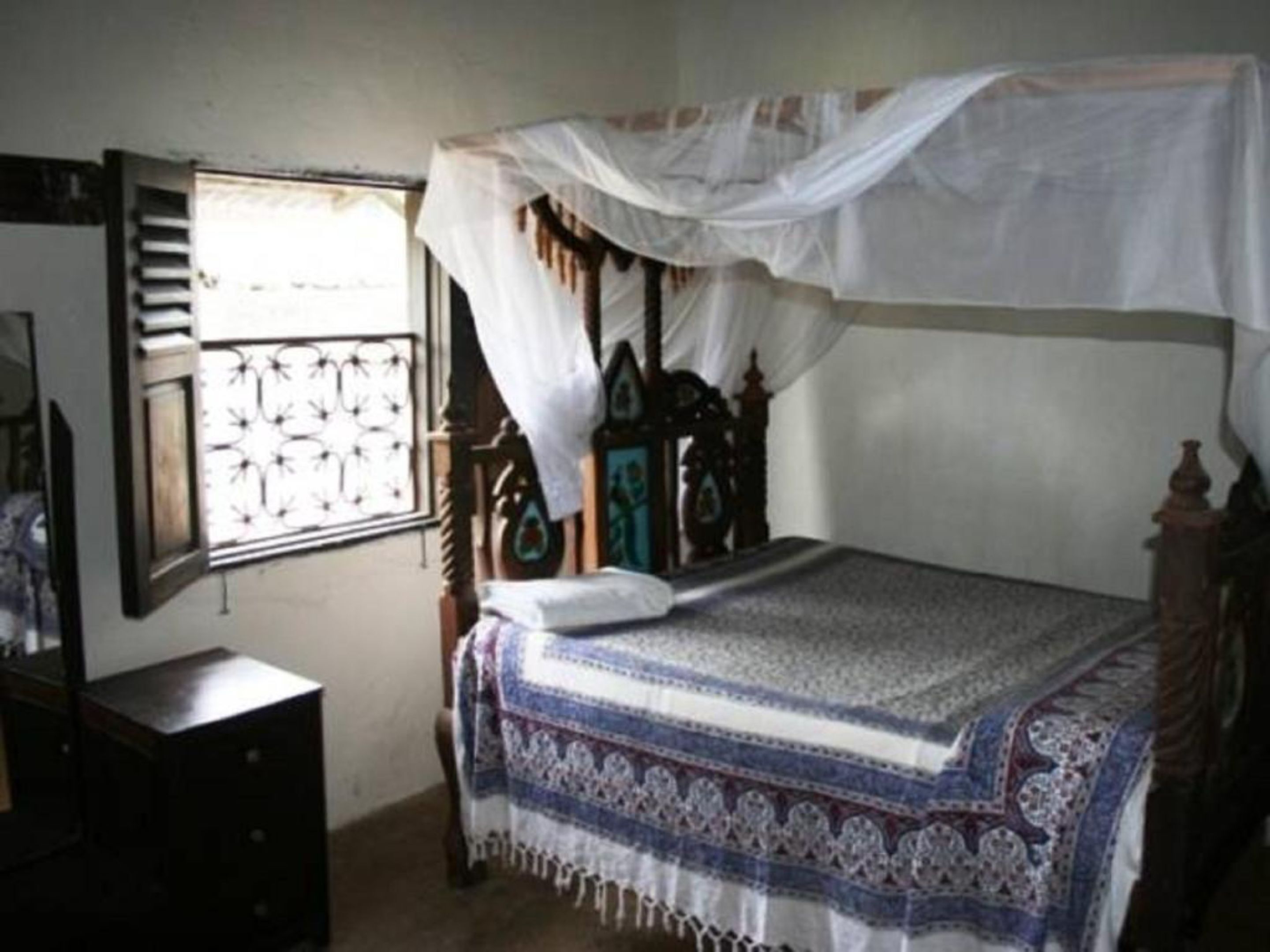 A bedroom with a bed under a mosquito net, a wooden headboard, a window with decorative grills, and a dresser.