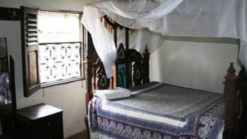 A bedroom with a bed under a mosquito net, a wooden headboard, a window with decorative grills, and a dresser.