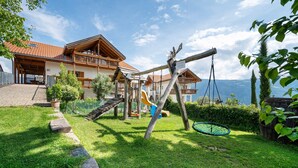 A chalet with a wooden deck, a swing set, and a view of the mountains. - Apartment 'Stettnerhof Penegal' with Mountain View, Terrace, Shared Garden & Wi-Fi (Völlan/Lana)