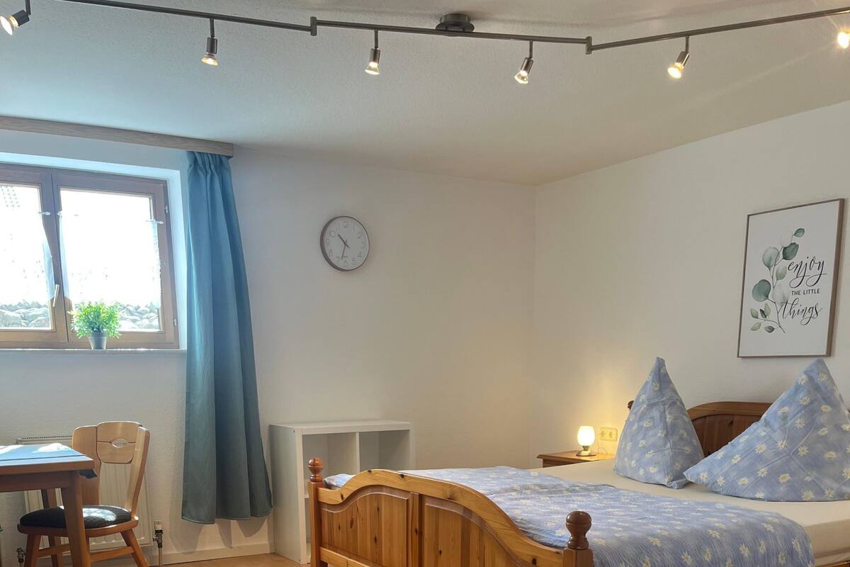 A bedroom with a wooden bed, a desk, and a chair. There is a window with curtains, a clock on the wall, and a framed picture.