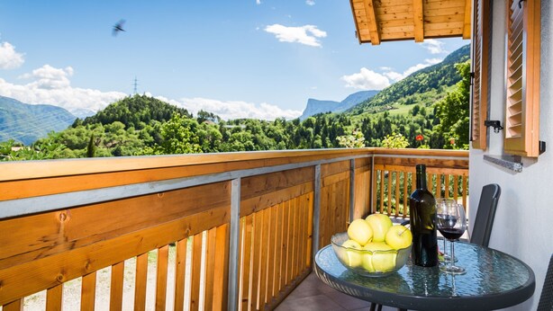 Ein Balkon mit Holzwertkasten, einem Tisch mit Flasche und Glas sowie einer Schüssel mit Äpfeln.