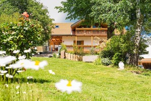 A two-story building with a balcony, surrounded by greenery and a grassy area.