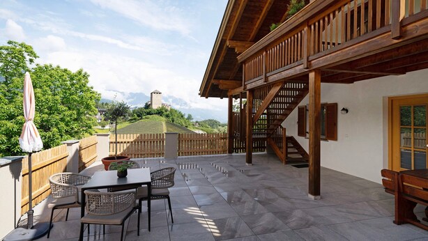 A patio with a table and chairs, a wooden deck, and a view of a hill with a tower. - Apartment 'Stettnerhof Mayenburg' with Mountain View, Terrace & Wi-Fi (Völlan/Lana)