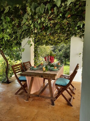 Un espace de restauration en plein air avec une table dressée pour quatre personnes, entourée de chaises en bois dotées de coussins bleus, le tout sous une canopée de feuillage verdoyant.