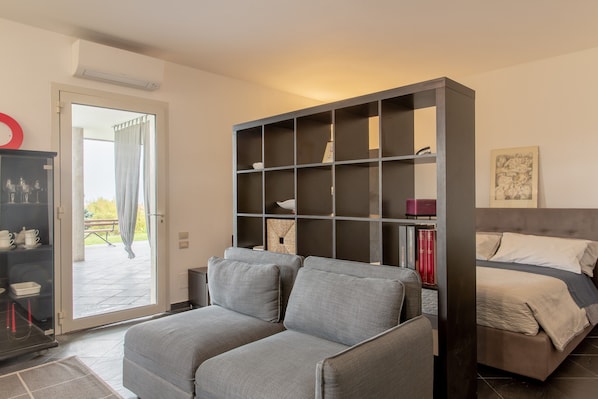 A modern bedroom with a grey sofa, a bed with a grey headboard, a bookshelf, and a sliding glass door leading to an outdoor area.