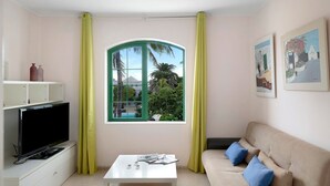 A living room with a TV, a white coffee table, a beige sofa with blue cushions, a window with a view of palm trees, and artwork on the wall. - Holiday Apartment 'Apartamento Rocas Negras' by the Beach with Pool View & Terraces (Puerto del carmen)