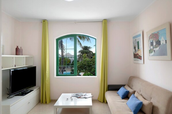 A living room with a TV, a white coffee table, a beige sofa with blue cushions, a window with a view of palm trees, and artwork on the wall. - Holiday Apartment 'Apartamento Rocas Negras' by the Beach with Pool View & Terraces (Puerto del carmen)